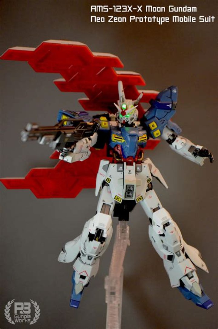 A Moon Gundam Gunpla kit looks at the camera while aiming it’s energy rifle at the lens and holds a beam saver to its side.